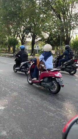 The video shows a side view of three individuals riding motorcycles on a paved road. The person in the foreground, on a red motorcycle, is wearing a white helmet and a blue headscarf, with a brown bag slung over their shoulder. They appear to be looking ahead and moving forward. Behind them, two other individuals are also riding motorcycles. The person to the left is wearing a blue helmet and a dark jacket. The person to the right is wearing a black helmet and a dark jacket. All three individuals are moving in the same direction on the road. The environment appears to be outdoors, with trees lining the road and some buildings visible in the background. The lighting suggests it might be daytime, possibly morning or late afternoon, with some sunlight filtering through the trees. The road surface is asphalt.