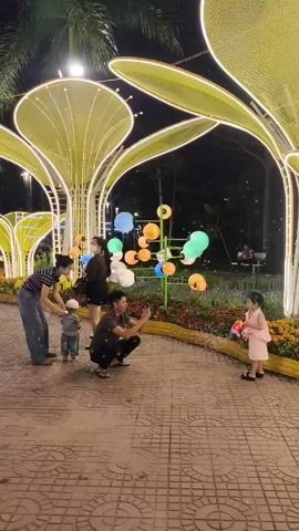 At night, a family is gathered around a walkway in a park decorated with brightly lit, abstract tree sculptures and colorful, decorative lanterns. A man kneels, taking a picture with his phone of a little girl holding a toy gun. A woman with a small child stands nearby. A second woman with a mask stands behind them. People can be seen walking on the pathway and sitting in the background. The ground is paved with patterned bricks. The night sky is dark.