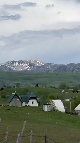 The scene is a rural landscape featuring a collection of small buildings with green roofs scattered across a lush, green field. In the background, a mountain range with snow-capped peaks rises against a cloudy sky. A winding road is visible through the landscape. A wooden fence runs along the bottom of the frame. The wind appears to be blowing.