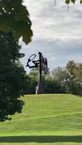 The video opens on a slightly blurry, distant view of a metal sculpture on a grassy hill. The sculpture is dark and appears to be abstract, with several intersecting geometric shapes. Lush green trees frame the shot on the left and top. The sky above is overcast with patches of blue. As the camera pans slightly, a hand enters the frame from the right, holding a red soda can. The can is in focus, obscuring part of the background. The video then continues to show the sculpture and the surrounding landscape, with the trees and sky still visible. The scene is set outdoors on a sunny day with scattered clouds, featuring a well-maintained lawn and mature trees.