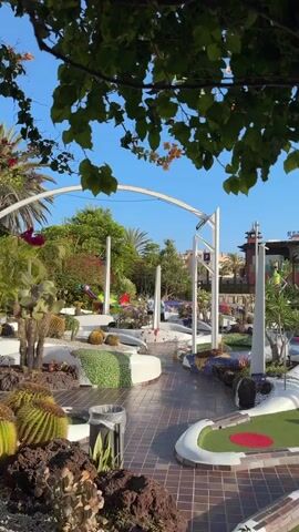 The video pans across a miniature golf course under a clear blue sky. The course features white, curved borders and green turf with red circular hole markers. Various cacti and other succulents are planted in dark rock beds surrounding the greens. In the background, a multi-story building with red tile roofs and balconies is visible, along with a tall, slender tower. Several people are scattered throughout the course, some sitting and others walking. The foreground is partially obscured by lush green leaves and branches of a tree.