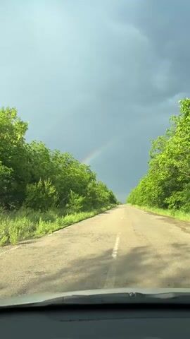 The video shows a view from a car driving down a paved road. The road is lined on both sides by lush green trees and grass. The sky is overcast with dark clouds, but a rainbow is visible in the distance. The camera is positioned in the front of the car, providing a first-person perspective of the drive. The road appears to be in a rural or natural setting.