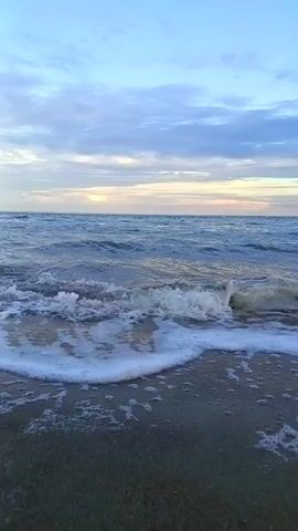 The video shows a view of a beach with gentle waves lapping at the shore. The sky is a soft blue with scattered clouds, and the sun appears to be setting or rising in the distance, casting a warm glow. In the foreground, the wet sand is visible, with foam from the waves creating patterns on the surface. As the video progresses, a young girl appears on the right side of the frame. She is wearing a blue cropped long-sleeved shirt and black shorts, and she is standing in the shallow water at the edge of the sea. The waves are washing around her ankles. The overall scene is serene and peaceful, capturing a moment at the beach.
