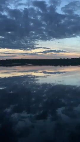 The video displays a serene body of water reflecting a cloudy sky at dusk. The horizon shows a dark silhouette of land. The clouds are a deep blue, interspersed with lighter streaks of yellow and pink from the setting sun. The water's surface is calm, creating a mirror image of the sky and land. The overall mood is peaceful and tranquil.