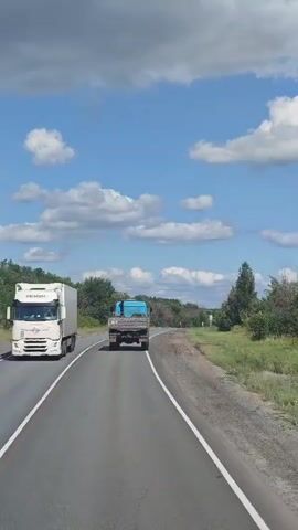 The video shows a view from a vehicle traveling on a paved road. A white semi-truck is visible on the left side of the road, partially in frame. Ahead, a blue truck is driving in the same direction. The road curves to the right. The landscape on either side of the road is green with trees and vegetation. The sky is blue with scattered white clouds.