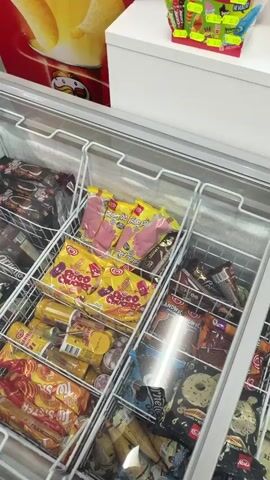 The video shows a freezer display case stocked with various ice cream treats.  Focus is given to several Frigo Pie brand foot-shaped popsicles in yellow packaging. Below them are Choco Choco brand popsicles with cartoon faces. To the side, there are ice cream sandwiches, and other assorted ice cream bars. The freezer is well-lit, and there is background music playing.