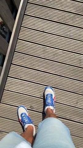 The video begins with a first-person view of someone standing on a wooden deck, looking down at their feet wearing blue and white sneakers. The camera then pans up and around, revealing a breathtaking panoramic view of a lush green forest and rolling hills under a clear blue sky. In the distance, a small town or village is visible nestled among the trees. The scene appears to be from an elevated viewpoint, possibly a lookout tower or a high vantage point on a mountain. The overall impression is one of natural beauty and tranquility.