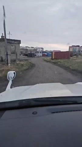 The video shows a view from inside a vehicle looking out at a dirt road. On the left side of the road are some buildings, one with broken windows. Further down the road, there are several shipping containers of various colors, including red, blue, and gray. A person is visible inside the vehicle, and the dashboard is in the foreground. The sky is overcast and gray, suggesting a cloudy or possibly rainy day. The environment appears to be an industrial or port area.