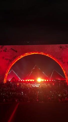 The video shows a large crowd of people gathered for an outdoor music event at night, with the Great Pyramids of Giza visible in the background. The pyramids and the entire performance area are illuminated with red lighting. There is a large, red, arch-shaped structure framing the view of the stage and the pyramids. The name "ZAMNA" is displayed on the stage. The crowd is densely packed and appears to be enjoying the music.