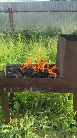 The video shows a close-up view of a rusty metal grill with burning charcoal and flames. The grill is outdoors, surrounded by green grass and tall weeds. A metal fence is visible in the background. The camera is static, and the focus is on the fire. There are no people or other objects visible in the frame.