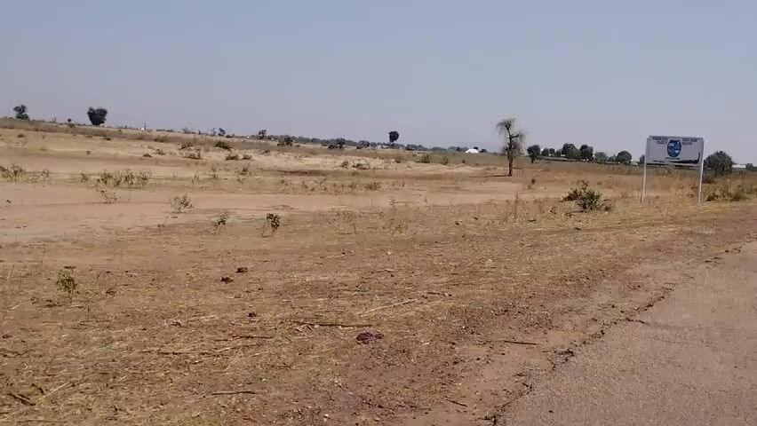 The video shows a mostly dry, dusty landscape with sparse vegetation under a bright blue sky. A road runs along the side of the landscape. A sign for "Kurami Girls College of AR Education" stands next to the road in the landscape. A car is seen driving down the road, carrying three people on top of it and an item wrapped in white fabric. The sound of wind is also present.