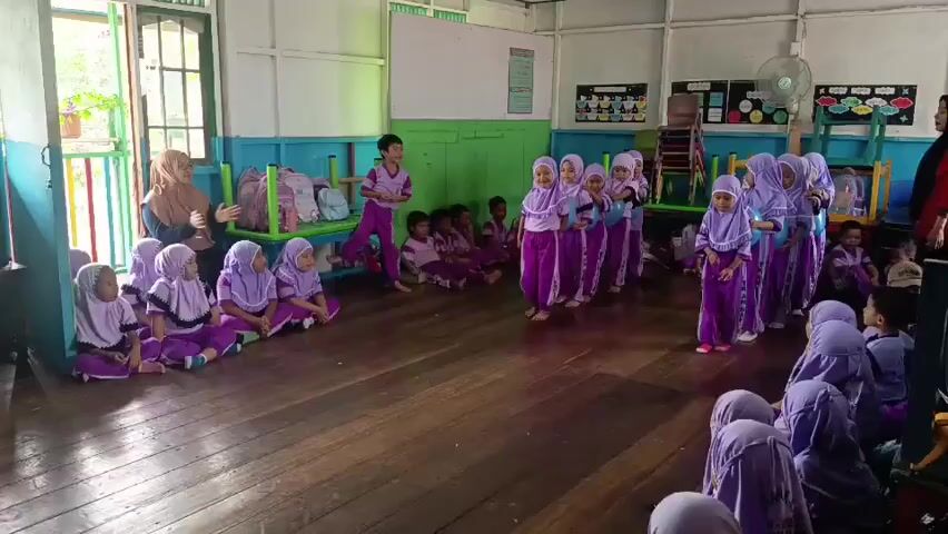 A group of young children, mostly girls wearing purple hijabs and matching purple outfits, are gathered in a wooden-floored room that appears to be a classroom. Some children are sitting cross-legged on the floor in two groups, facing each other. Another group of girls, also in purple outfits and hijabs, are standing in a line in the center of the room, holding blue balloons. One girl in the standing line is slightly ahead of the others. A few other children, and at least two adults, are present. One adult, a woman in a brown hijab and a long skirt, is sitting on a chair to the left. Another adult, a woman in a beige hijab and a dark top, is standing on the far left of the frame, facing the children. A young boy in a purple outfit is standing near the back of the room. The room has green and blue painted walls, windows, and various classroom items visible in the background, including chairs, backpacks, and a whiteboard. The children appear to be engaged in some sort of activity, possibly a game or performance. The overall atmosphere seems to be one of a children's educational setting.