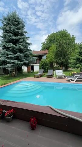 The camera pans right from a swimming pool to a covered patio. The patio has a wooden ceiling and walls. A large television is mounted on a wooden wall, displaying a video game. Below the TV are various electronics and a mini-fridge. A hammock is strung across the patio. A table with drinks and a gaming controller is visible. A person is sitting at the table, playing the video game. Outside the patio, a lush green landscape with trees and bushes can be seen under a blue sky.