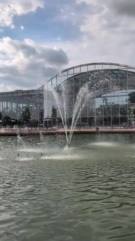 A wide shot shows a body of water with several fountains spraying water into the air. Behind the water is a large building with a curved glass roof and facade. Trees and outdoor seating are visible on the walkway in front of the building. The sky is cloudy.