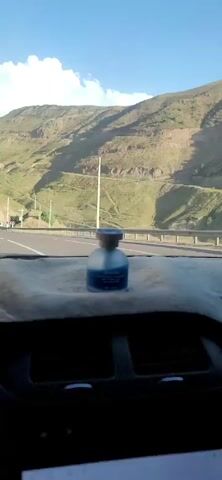 The video shows a view from inside a car driving on a highway. The dashboard is visible in the foreground with an air freshener on it. The highway is paved and has a guardrail on the right side. The landscape outside is mountainous and appears to be dry with some vegetation. There are power lines and poles visible along the side of the road. The sky is blue with some clouds. There are other cars on the road ahead. The camera is moving forward, indicating the car is in motion.