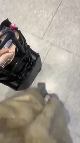 A person is pulling a black rolling suitcase across a tiled floor. The suitcase appears to contain a pair of black high-heeled shoes and other items. The person's hand is visible on the handle of the suitcase. Only the lower part of their body is visible, showing pants with a speckled pattern, white shoes, and tan fur. The floor is composed of large, light gray tiles.
