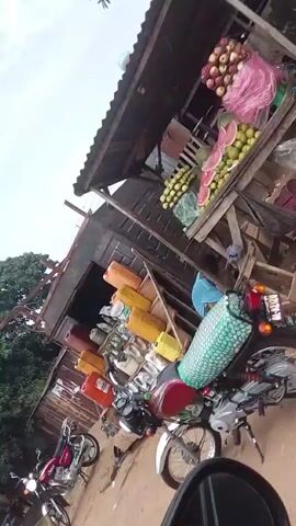 The video begins with a close-up of a red motorcycle parked next to a dark-colored vehicle. The camera then pans to the right, showing a corrugated metal building with a white tarp extension. Beyond that, trees and a cloudy sky are visible. The scene appears to be an outdoor area, possibly a roadside or a market.

As the video progresses, the camera moves, showing more of the surrounding environment. There are more buildings, some with multiple stories and windows. Several motorcycles are seen parked or moving. People are visible in the background, though their features are not clear. Bags of what appear to be goods are stacked near some of the buildings. The ground is a dusty, reddish-brown color, suggesting a dirt or unpaved road. The camera angle is often tilted, giving a dynamic but somewhat disorienting perspective of the location. The overall impression is that of a busy, possibly rural or semi-urban environment with evidence of commerce and transportation.