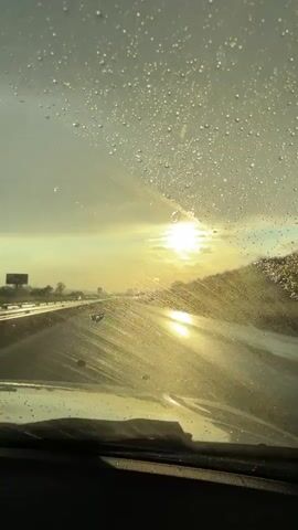 A view from inside a car driving on a highway during or after rain. The sun is shining brightly through a cloudy sky, reflecting off the wet road. Water droplets cover the windshield, which is being cleared by wipers.