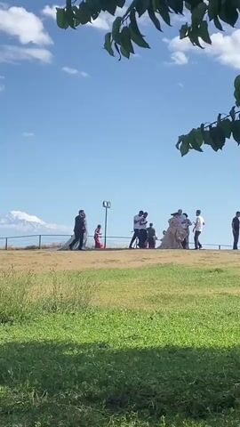 A group of people are gathered on a grassy hill under a blue sky with scattered clouds. In the background, a snow-capped mountain is visible. Several individuals appear to be involved in a wedding ceremony or photoshoot. A man in a suit and a woman in a white wedding dress are positioned towards the left of the frame, with a long train of the dress trailing behind them. Other people, some holding cameras, are observing and participating in the event. The foreground is dominated by lush green grass. The lighting suggests it is daytime, likely sunny.