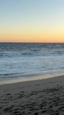This is a vertical video taken on a beach at sunset. The sky is a gradient of pale blue to orange. The ocean is visible with gentle waves rolling onto the shore. The beach is sandy with footprints visible in the foreground. A faint silhouette of a mountain range is visible on the horizon. There are no people or objects clearly visible in the frame. The video shows the sun slowly descending behind the mountains.