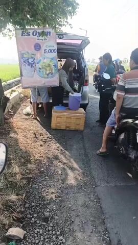 A roadside vendor is serving drinks from the back of an open SUV. The vendor, a woman wearing a headscarf, is interacting with a customer. A sign displays "Es Oyon" and a price of "5.000". Several people on motorcycles are present, suggesting a busy transaction point. The scene takes place outdoors, with a paved road, a grassy area, and a rice field visible in the background under a bright sky.