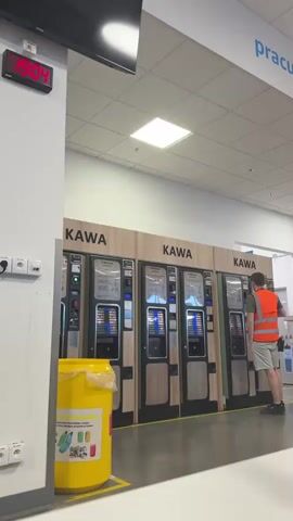 This video shows a row of three coffee vending machines labeled "KAWA" in what appears to be a public space, possibly a store or service area in Chociule, Poland. A digital clock on the wall displays the time as 19:04. A person wearing an orange safety vest and shorts is standing in front of the rightmost vending machine, interacting with it. Another person is partially visible behind them. A large yellow recycling bin with images of bottles and cans is situated to the left of the vending machines. The overall lighting suggests it is daytime, but the time of 19:04 implies it is late afternoon or early evening. The scene is indoors, with a tiled floor and a suspended ceiling with fluorescent lighting. The machines appear to be operational, with illuminated displays. Text visible on the vending machines includes "KAWA" (Polish for coffee) and instructions or advertisements, although the text is too small to read clearly. The setting appears clean and organized.