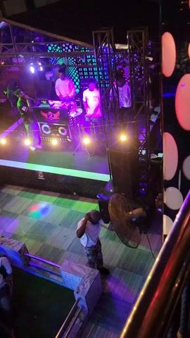 The video captures a live music performance on an elevated stage in an indoor venue, likely a club or bar, at night in Awada, Nigeria. Multiple men are on stage: one is singing into a microphone, another is playing bongo-style drums, and a third is positioned near a DJ console or mixer. The stage is dynamically lit with vibrant, changing colors including green, pink, blue, and purple, complemented by warm yellow incandescent bulbs. The background features decorative panels with geometric and circular patterns, supported by metal trussing. A speaker box on the stage displays the text "STEP 99.4" and a circular logo. The drummer wears a white t-shirt with "999" visible. Below the stage, a man is seen walking past a large floor fan.