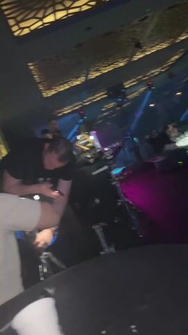 The video depicts a scene inside a crowded bar or club with a lively atmosphere. Several individuals are visible, some standing and others seated. The lighting is colorful and dynamic, with a mix of purple, green, and red hues that contribute to the club's ambiance. The camera moves around, focusing on people and objects within the space. Red and blue cube-shaped seats can be seen. The ceiling features a geometric design, adding to the visual appeal of the club. The sound of music is audible, enhancing the lively environment.