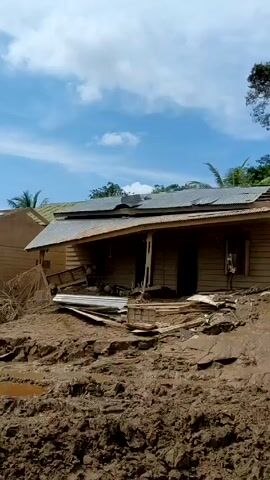 The video shows the aftermath of a natural disaster, likely a mudslide or flood, in a tropical region. Wooden houses are heavily damaged or destroyed, with mud and debris covering the landscape. There are palm trees present. The ground is covered in thick mud and debris. In some shots, people can be seen walking around, assessing the damage and potentially clearing debris. One man is wearing a vest with the word "BAZNAS" on it. The sky is visible with varying degrees of cloud cover, and the weather appears to be sunny. The overall scene conveys a sense of destruction and the need for recovery efforts.