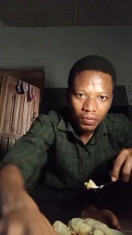 A young man is sitting cross-legged in a dimly lit room, wearing a dark green plaid shirt and black pants. He's holding a fork with food on it, eating it, and then reacts with shock, opening his mouth wide and grabbing his head. There is a plate of food next to him on the floor, covered in a plastic bag.  Behind him is a wooden door slightly ajar with some clothes hanging on it. The ceiling is a light grey.