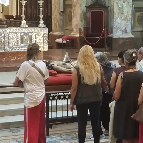 The video shows a crowd of people inside what appears to be a large, ornate church. At the front of the room, a group is gathered around a table on which lies a statue of a figure resembling the deceased Christ. The people are observing the statue. A man wearing a hard hat and safety vest moves through the crowd. The church interior features large marble pillars, religious paintings, and a decorative altar. The atmosphere is quiet and reverent, with the sound of a choir in the background.