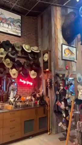The camera slowly pans across the interior of what appears to be a Western wear store. The room features exposed brick walls, a decorative ceiling, and wooden shelving and cabinets. A collection of cowboy hats is displayed on the wall above a row of cabinets, illuminated by a neon sign that reads "Pretty Gang" and "Swagger". To the left, a tall bookshelf showcases rows of cowboy boots, along with a few bags and other items. Adjacent to this is a red refrigerator-style container adorned with polaroid photos. The walls are decorated with mounted animal heads, paintings, and other Western-themed artifacts.