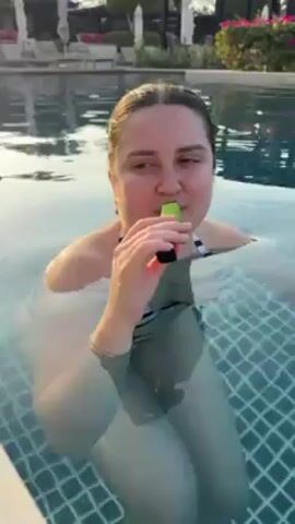 A woman with light skin, wearing a black-and-white striped swimsuit, is standing in a swimming pool with blue tiles. She is holding a black and lime green vape pen and inhales, then exhales a large puff of white smoke. The background shows lounge chairs with white umbrellas, a pink-flowering bush, and trees.