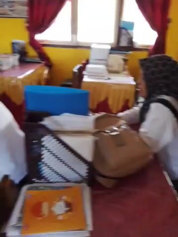 The video shows a classroom setting with yellow walls and red curtains. Several tables are covered with red and gold cloths. People are seated at the tables, some working on laptops and papers. Two individuals, possibly students or teachers, are standing and interacting near a table. The overall atmosphere appears to be one of an educational institution.