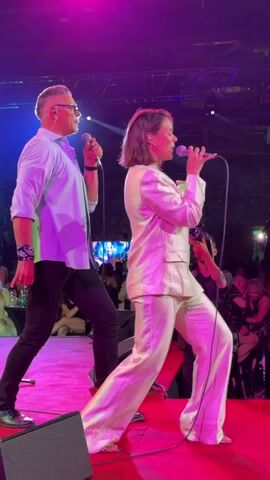 This is a video of two singers on a stage, performing in front of an audience. The male singer on the left is wearing a light blue shirt, dark pants, and glasses, and is holding a microphone. The female singer on the right is wearing a white suit and also holding a microphone. The stage is lit with colorful spotlights, including blue, green, and pink. There are speakers on the stage and some audience members can be seen in the background. The singers are both in motion, as if dancing or gesturing while they sing. The video is from a concert or live performance.
