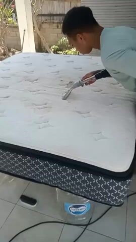 A young man in a light blue t-shirt is vacuuming a mattress outdoors. The mattress is white with a grey and black patterned border and is propped up on a large clear plastic water jug. The man is holding a vacuum cleaner attachment with his right hand and is pressing it onto the mattress. The mattress appears to have some stains on it. The background shows a white wall and some greenery.