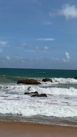 A calm, sunny day at the beach with gentle waves breaking on the shore. Several large rocks are visible in the shallow water, with white foam from the waves washing over them. The ocean is a deep turquoise color, and the sky is a clear blue with a few scattered white clouds. The sand in the foreground is a light brown color. No people are visible in the video.