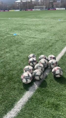 The video shows a soccer field with a pile of soccer balls at one edge of the field. The balls are white and black. The field is green artificial turf. The edge of the field is marked by a white line. There are blue cones scattered on the field. In the background are buildings, trees, a mountain, and a cloudy sky. A person wearing a black shoe is standing near the pile of soccer balls.