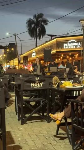 This is a video of an outdoor dining area at night. In the background, a building with the sign "PATAGONIA BREWING CO." is visible, illuminated with yellow lights. A live band is playing on a stage in front of the building, with a singer at the microphone. In the foreground, there are tables and chairs with the branding "HOLY BREWERY" on the chairbacks. Some patrons are seated at the tables, eating and drinking. The overall atmosphere suggests a lively, casual evening at a restaurant or bar.