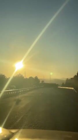 The video shows a car's perspective driving on a highway towards the sun. The sun is low in the sky, shining brightly, causing glare. Other cars are visible on the road, and there are power lines in the background. The overall tone is bright and sunny.