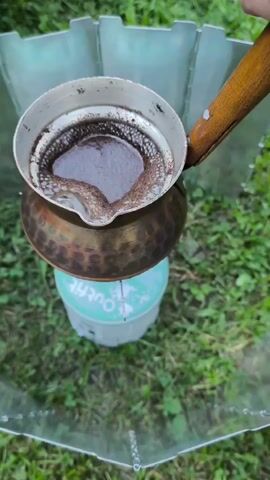 The video shows a close-up, outdoor shot of Turkish coffee being brewed. A copper cezve, filled with dark liquid and foam, sits on a small, green camping stove. The stove is branded with the word "Outfit". The cezve has a wooden handle. A silver wind guard is positioned around the stove and cezve. The background consists of green grass and foliage. In the later frames, a hand reaches in to grasp the handle of the cezve and lifts it off the stove, which has a visible flame.