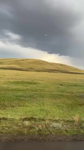 The media file captures a view from a moving vehicle, likely a bus or car, traveling through a rural landscape in Kazakhstan, specifically near Bürkitti. The scene is dominated by rolling, grassy hills under a dramatic, overcast sky. Dark, heavy clouds suggest impending or ongoing rain, creating a somber and slightly moody atmosphere. The ground is a mix of vibrant green grass and drier, yellowish patches, indicating a semi-arid environment.

A road, appearing wet and possibly recently rained on, runs alongside the grassy terrain. In the distance, a road sign is visible, its specific details unclear due to the distance and resolution. Further along the road, a concrete overpass or bridge is prominently featured. The bridge is supported by sturdy concrete pillars and has a metal railing along its edge. The embankment leading up to the bridge is reinforced with concrete blocks. The overall impression is one of a vast, open landscape with signs of human infrastructure adapting to the natural environment. The time of day appears to be late afternoon or early evening, given the muted light and the intensity of the clouds. There are no people visible in the media file, but the presence of the road and bridge implies human activity and passage. The video pans slightly, offering a continuous view of the changing landscape as the vehicle moves forward.