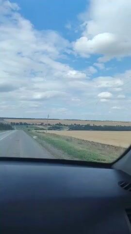 The video shows a view from the interior of a moving car. The perspective is looking forward through the windshield. Outside the car, a two-lane paved road stretches into the distance. The road is bordered by a grassy shoulder and fields of dry, harvested crops. In the distance, there are trees, some buildings, and utility poles. The sky is blue with scattered white clouds. The image is slightly blurry, suggesting motion.