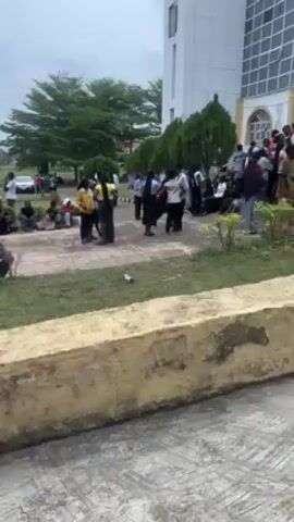The video shows a gathering of people outdoors on a cloudy day. In the foreground, a person wearing a purple shirt and dark jeans with slippers walks from left to right. As the camera pans upwards, it reveals a group of people, mostly dressed in what appears to be school uniforms, sitting on a low grassy area next to a walkway. Some individuals are standing, and a few are holding umbrellas for shade. In the background, there is a building, possibly a school or university, and utility poles stretch along the road. The overall impression is of a school or campus setting with students gathered for an event or activity.