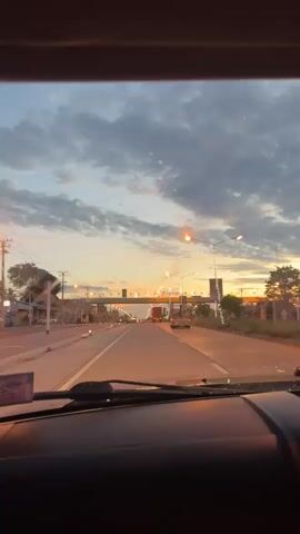 The video shows a point-of-view shot from inside a vehicle driving on a road at dusk. The sky is filled with dramatic clouds in shades of gray, orange, and blue, indicating either sunrise or sunset. Streetlights and traffic lights illuminate the road ahead, casting a warm glow. Several vehicles are visible on the road, including cars and trucks. A large overhead sign with decorative lights and text in an unknown language spans across the road in the distance. The road has multiple lanes and white dividing lines. The interior of the vehicle, including the dashboard and windshield wipers, is partially visible in the foreground. The overall scene conveys a sense of travel and movement during a transitional time of day.