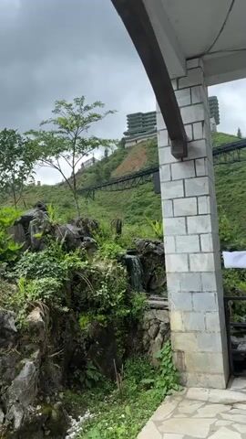 Here is a description of the video:
A woman in a white robe walks from behind a stone pillar onto a stone patio, and spreads her arms. She is wearing a red dress underneath. She is standing near a black railing. Behind the railing is a flowing stream. The background features a green hillside with a hotel on top, and a crane descending the hillside. The sky is gray.