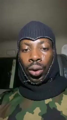 A man is seated inside of a car. He is wearing a black knit balaclava and a camouflage shirt. He looks at the camera and purses his lips.