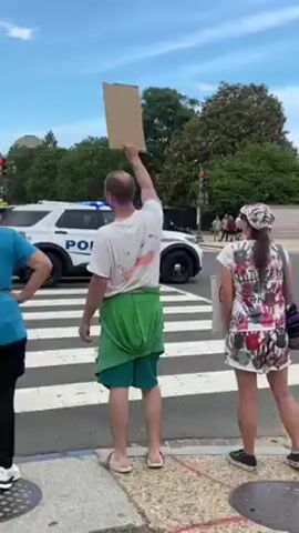 This media file depicts an outdoor scene of a protest or demonstration at a crosswalk in an urban environment on a clear, sunny day. The sky is bright blue with scattered white clouds.

In the foreground, three individuals are seen from behind. A man in the center, wearing a white t-shirt with some colored stains, green shorts, and light-colored flip-flops, holds a blank rectangular cardboard sign high above his head with his right arm. To his right, a woman in a colorful, patterned t-shirt, white shorts, and a matching cap stands, also appearing to hold a sign or another object, which is partially obscured. The top of another person's head and shoulder are visible to the man's left.

The street features a white-striped crosswalk. Beyond it, a white police car with "POLICE" visibly emblazoned on its side is parked or moving slowly. Further down the street, a white pickup truck and a smaller utility vehicle are visible. Numerous other people can be seen walking on the sidewalks and in the distance, indicating a public gathering. Green trees line the background, and a dome-shaped building is faintly visible on the horizon, suggesting a prominent city landmark.

The audio accompanying the video includes a woman's voice speaking passionately, with the audible phrase "who is a known child rapist," providing critical context to the subject matter of the protest. The scene captures a moment of public expression and activism.