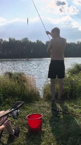 A young male with blonde hair, shirtless and wearing black shorts, stands facing a body of water, holding a fishing rod. The fishing line extends over the water, with a lure visible. To his left, a red bucket sits on the grassy bank. Another fishing rod lies on the ground near the bucket. In the foreground, a person's legs and arms are visible, seated in a chair and holding a fishing rod. The environment is a natural setting with green grass and trees lining the opposite bank of the water. The sky is bright with clouds, indicating daytime. The video captures a peaceful moment of fishing.