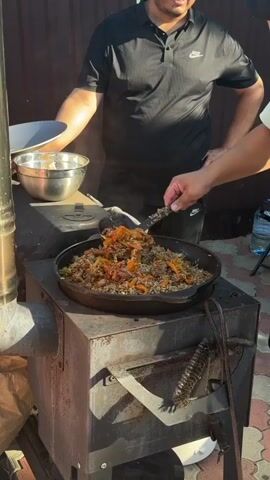 Two men are outdoors by a metal stove. One man, wearing a black t-shirt and black pants, stands to the left holding a silver bowl. The other man, wearing a black t-shirt and black pants, is on the right, using a black slotted spoon to stir a large black pot filled with rice, meat, and carrots. The pot is on top of the stove. Steam is rising from the pot. The background shows a brown fence and green leaves. The ground is paved with brick-like stones.
